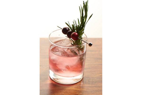 A glass of pink cocktail with ice, garnished with a rosemary sprig and skewered cranberries, sits on a wooden surface—perfect for celebrating Christmas in July.