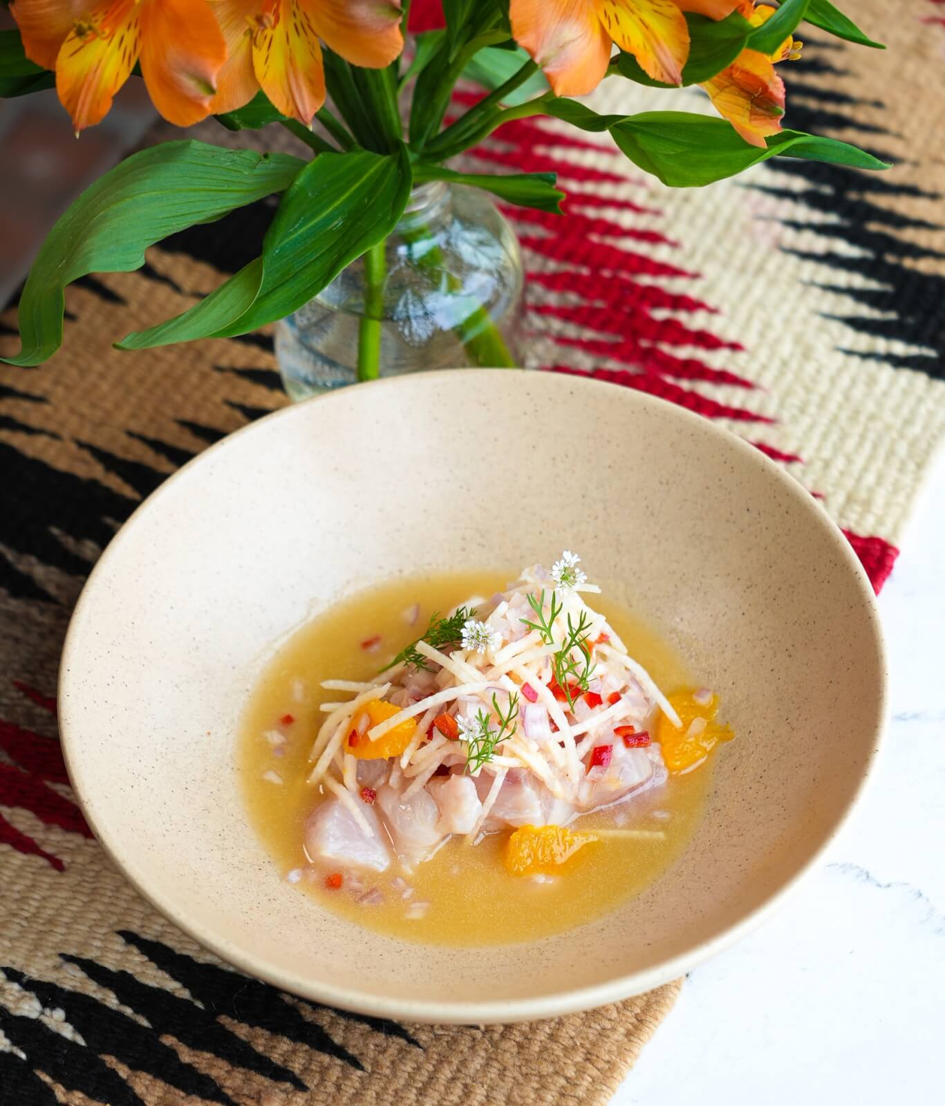 3 New Louisville Restaurants (+2 New Menus!) to Try This Month - 4 A beige bowl of ceviche with diced fish, shredded vegetables, and herbs in a citrus broth sits on a woven mat beside a vase of orange flowers.