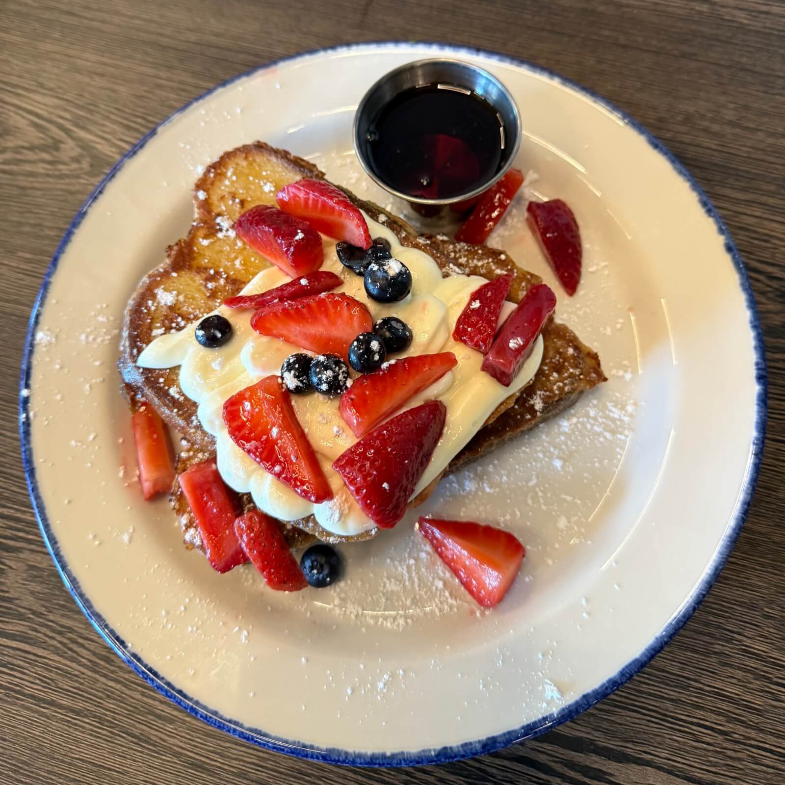 3 New Louisville Restaurants (+2 New Menus!) to Try This Month - 1 French toast topped with whipped cream, sliced strawberries, blueberries, and powdered sugar, served with a small cup of syrup on a white plate—a delicious brunch favorite found on many Louisville restaurant menus.