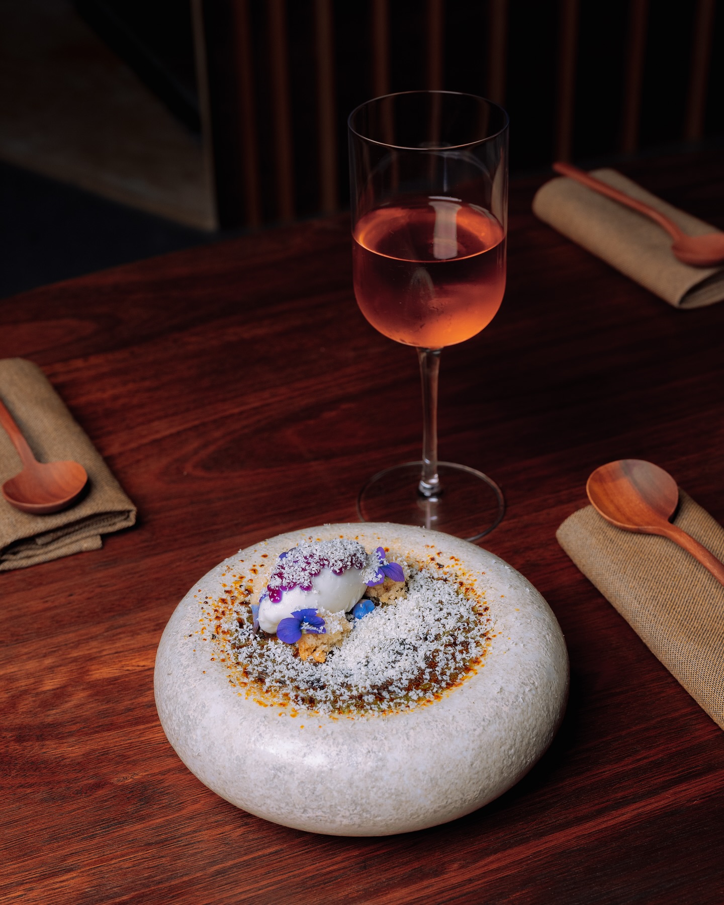 3 New Louisville Restaurants (+2 New Menus!) to Try This Month - 2 A stone plate with a gourmet dessert topped with white powder and purple flowers, next to a glass of rosé wine on a wooden table set with wooden spoons and brown napkins at one of the new Louisville restaurants.