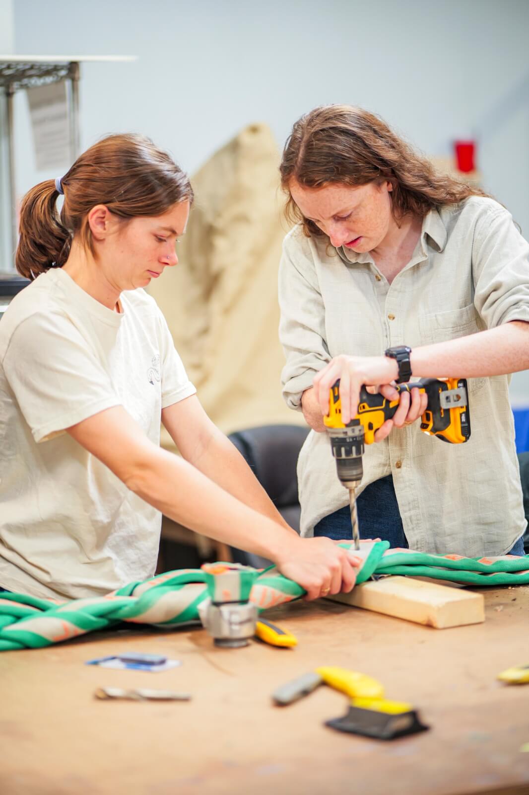 Two women use a power drill to secure a thick braided rope onto a wooden board at a workbench with various tools.