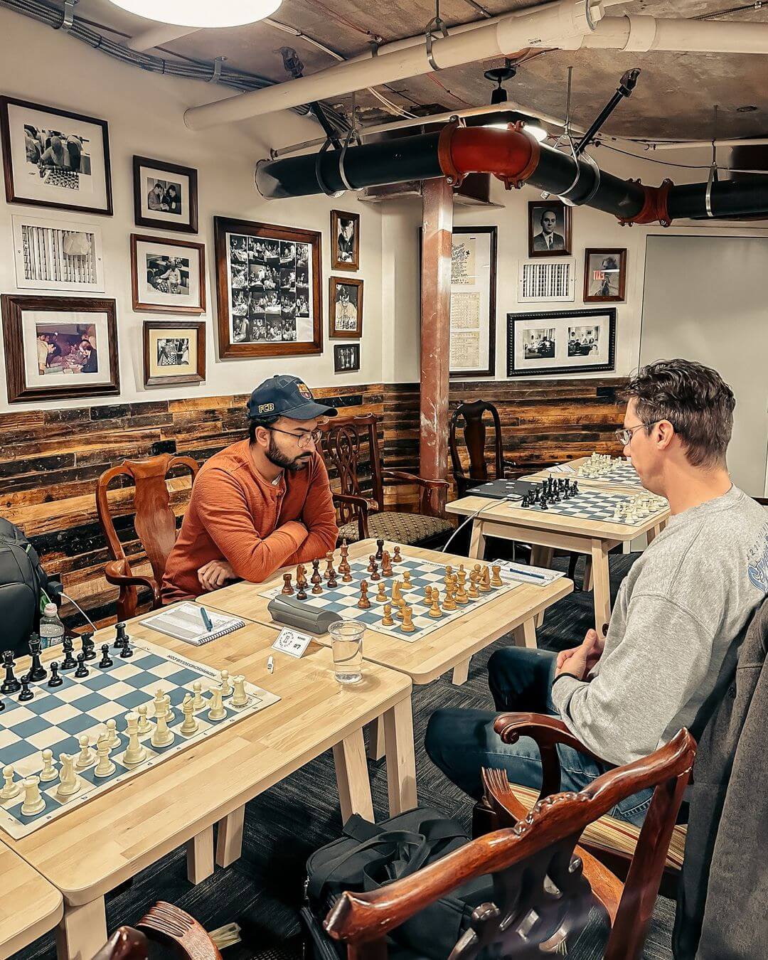 Two men are playing chess at a small table in a cozy, wood-paneled room decorated with framed photos and chess memorabilia.