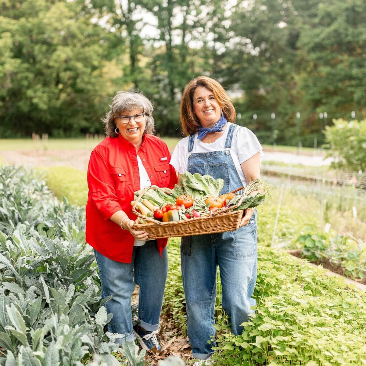 Two women stand in a garden, holding a large basket filled with freshly harvested vegetables.