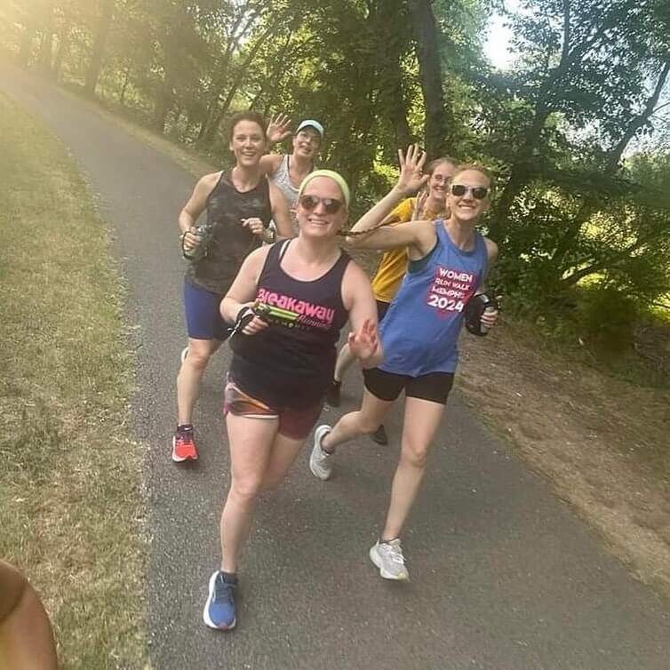 Five women jog and smile on a paved park path, wearing athletic gear and sunglasses, with trees and sunlight in the background.