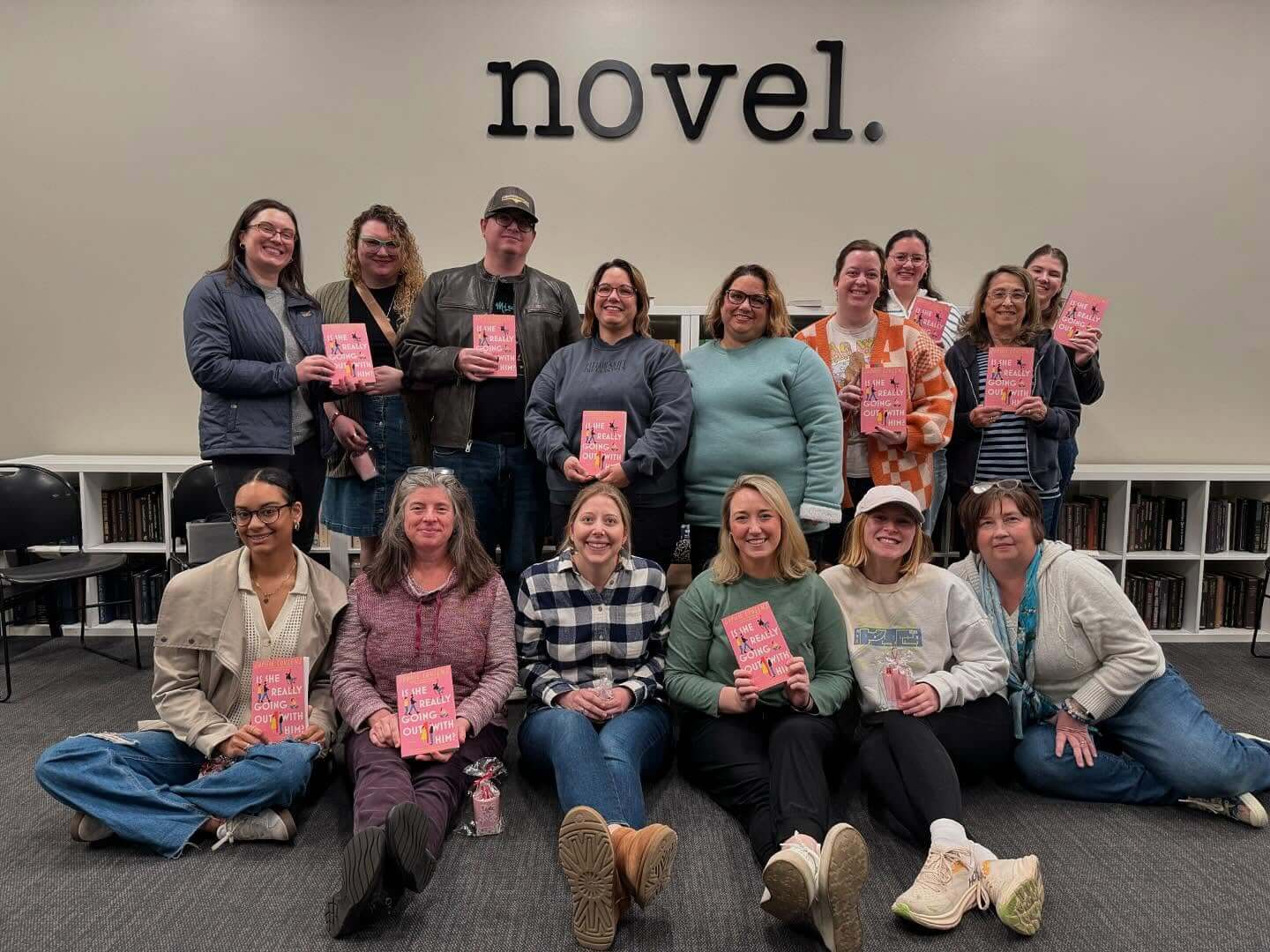 A group of Grown-Ups pose indoors, each holding a copy of the same book, under a wall sign that reads "novel," showing off their book club pick.