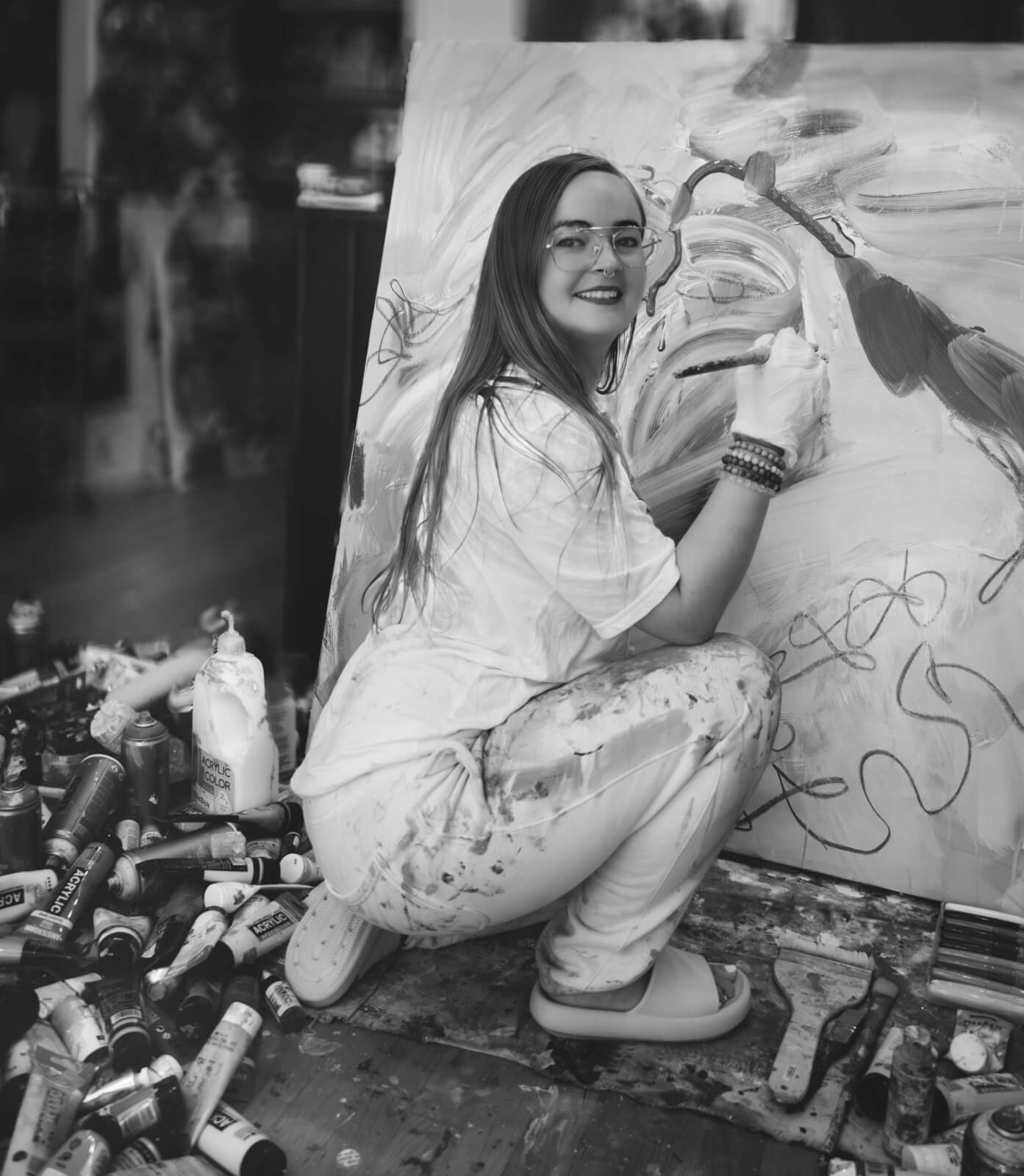 A woman in paint-splattered clothes crouches and smiles while painting an abstract artwork on a large canvas, surrounded by various art supplies.