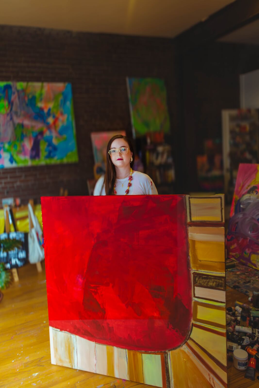 A woman stands in an art studio, holding a large canvas with a bold red abstract painting. Various colorful artworks and supplies are visible in the background.