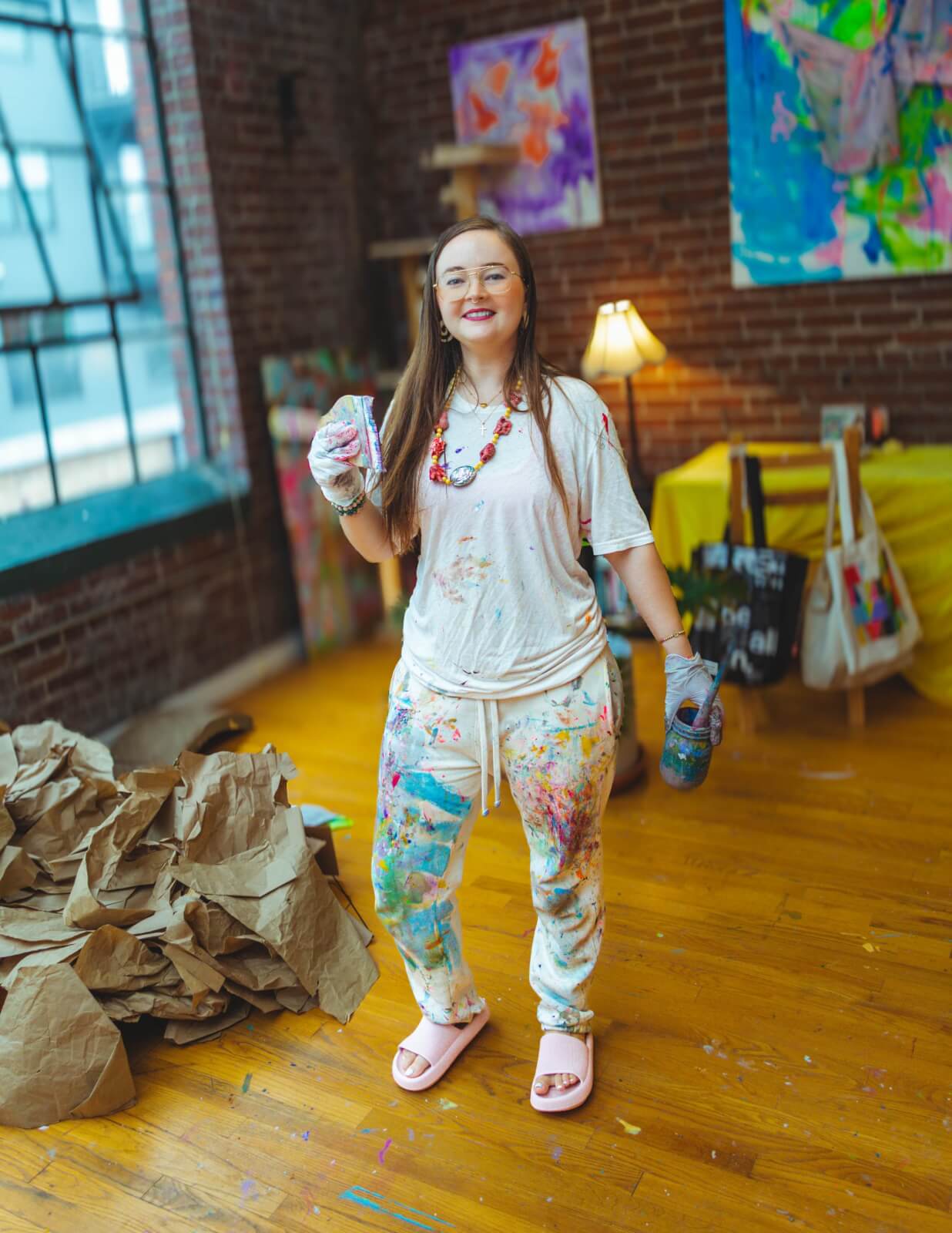 Alexandra Baker, in paint-splattered clothes, stands in an art studio holding a paint bottle, surrounded by paintings from her FACES series, crumpled paper, and art supplies.