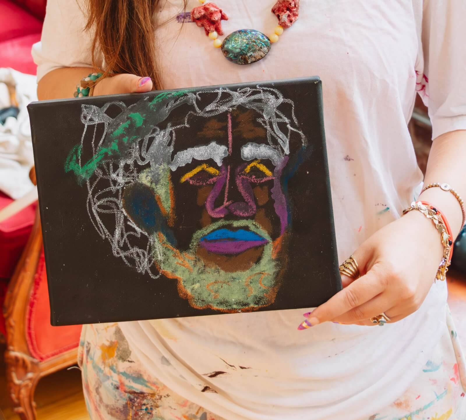 Person holding a canvas—a colorful, abstract portrait sketched in chalk or pastel on a black background. The person wears a white shirt with paint stains.