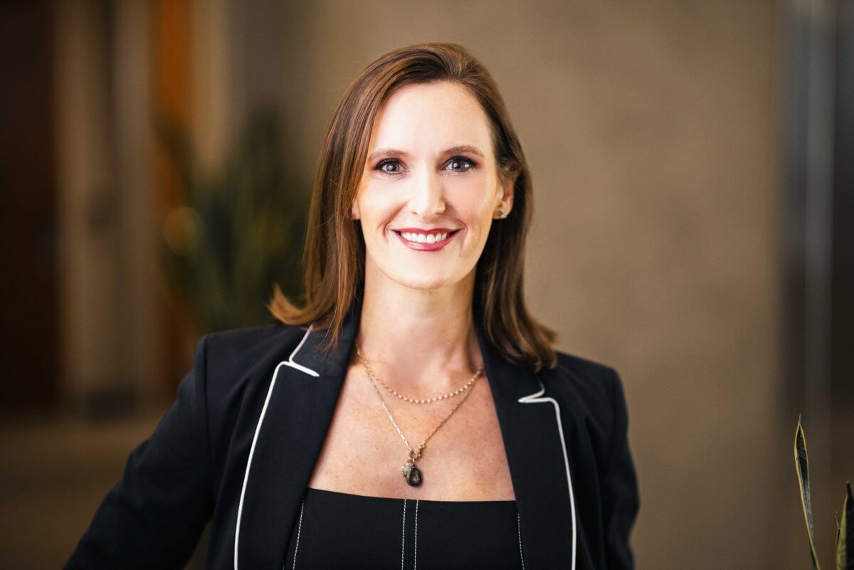 Carey Campbell, featured in FACES, is a woman with straight brown hair, wearing a black blazer with white piping, a black top, and layered necklaces as she smiles at the camera indoors.