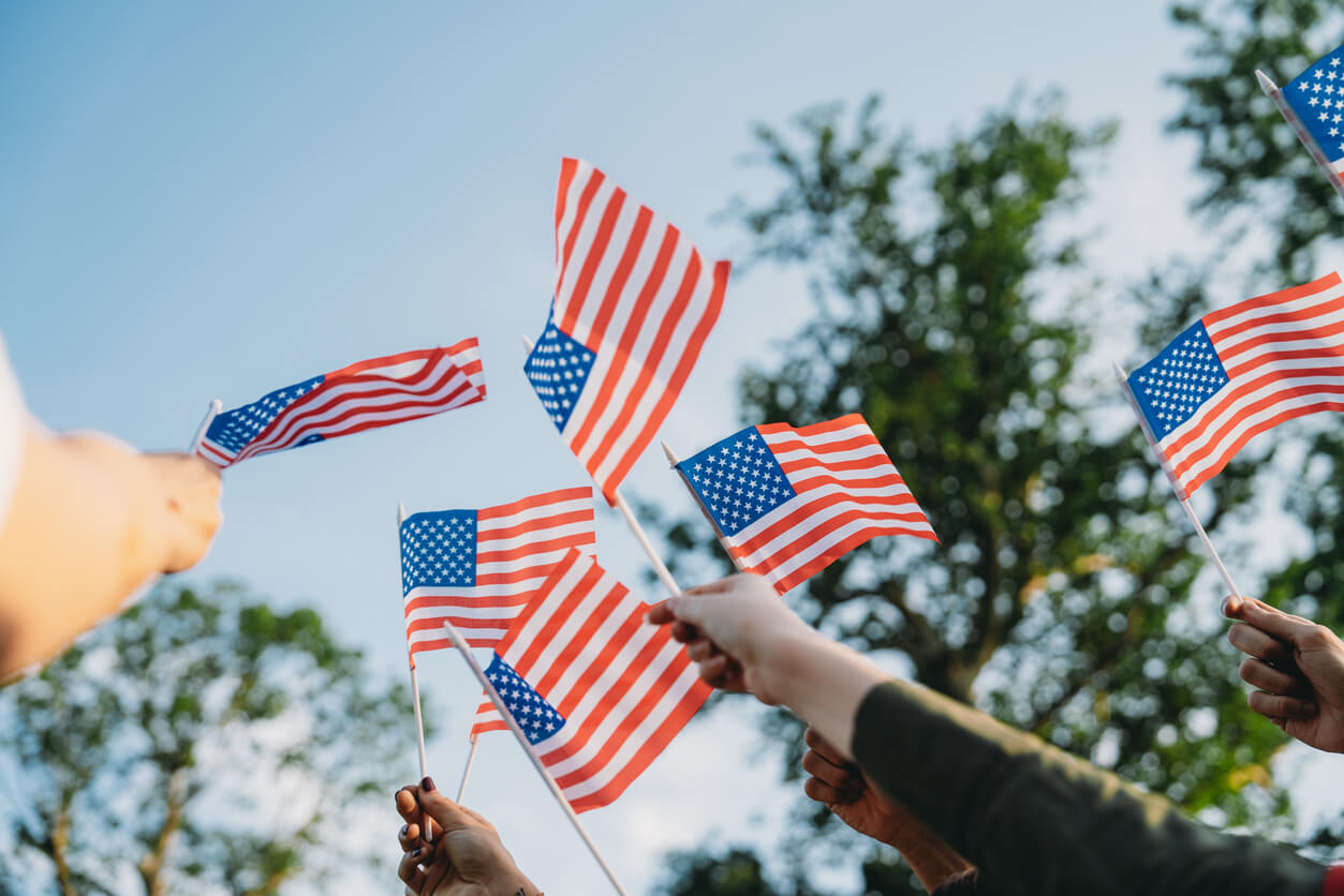Several hands hold small American flags outdoors against a blue sky with green trees in the background.