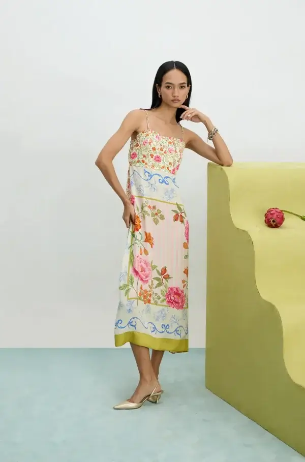 A woman stands indoors in a luxury floral midi dress with spaghetti straps and pointed-toe heels, leaning one arm on a curved yellow structure with a single pink flower—capturing the bold Memphis vibe of July 2025.