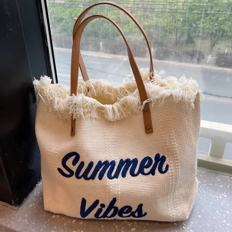 A beige woven tote bag with frayed edges and brown handles sits on a windowsill. "Summer Vibes" is printed on the front in blue lettering—one of Memphis’s hot finds for summer 2025.