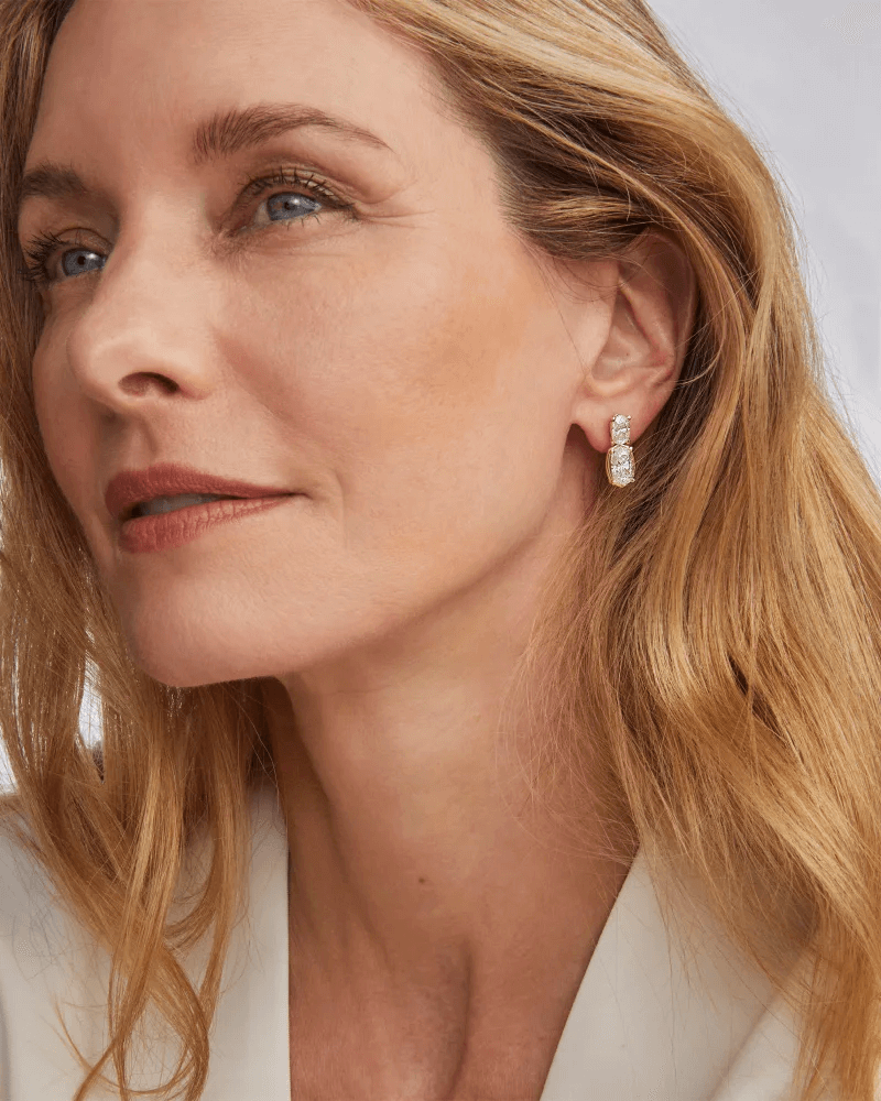 A woman with light brown hair wearing a white top and luxurious diamond drop earrings looks slightly upward with a neutral expression, capturing an elegant moment in Memphis, July 2025.