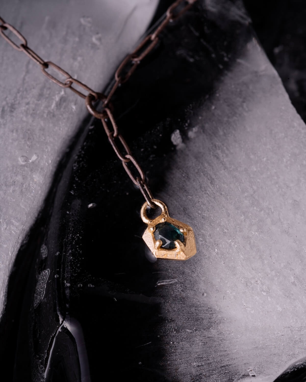 A close-up of a gold pendant with a dark gemstone hanging from a bronze chain, placed on a textured black and white surface, exuding the luxurious Memphis vibe of July 2025.