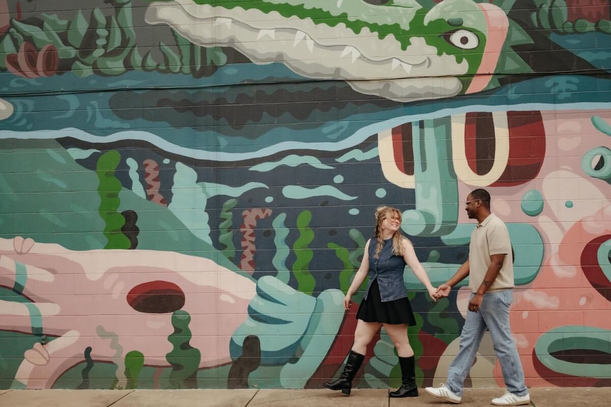 A woman and a man holding hands walk past a colorful memphis mural featuring an abstract crocodile and aquatic plants.