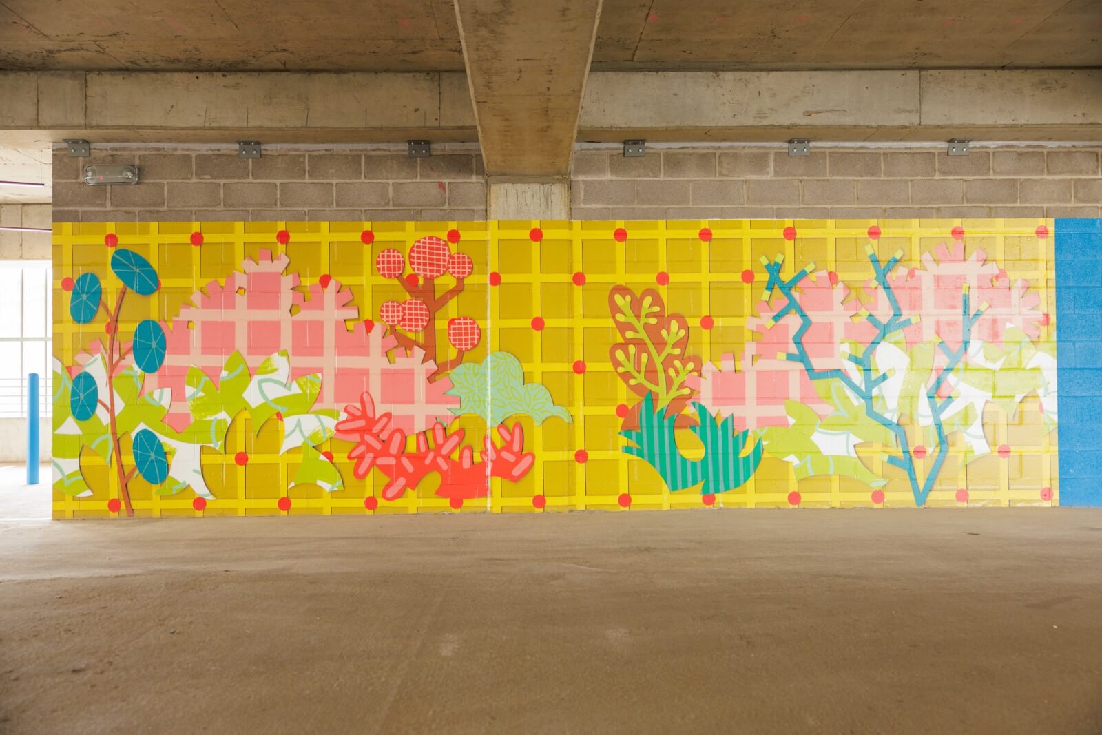 A colorful memphis murals-inspired mural with abstract plants and foliage in pink, green, and blue tones is painted on a yellow grid-patterned wall in a concrete parking structure.