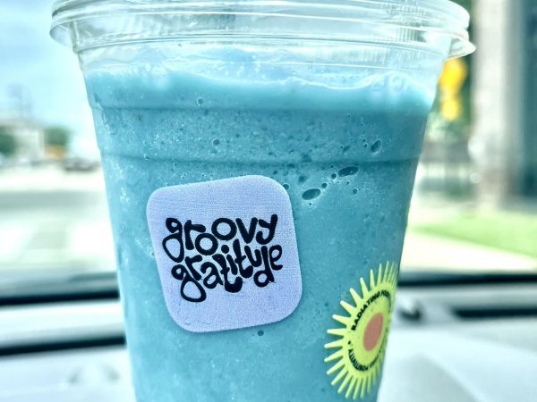 A clear plastic cup with a blue smoothie, a lid, and a "groovy gratitude" sticker on the front sits on a car dashboard—perfect refreshment while exploring new restaurants in Memphis.