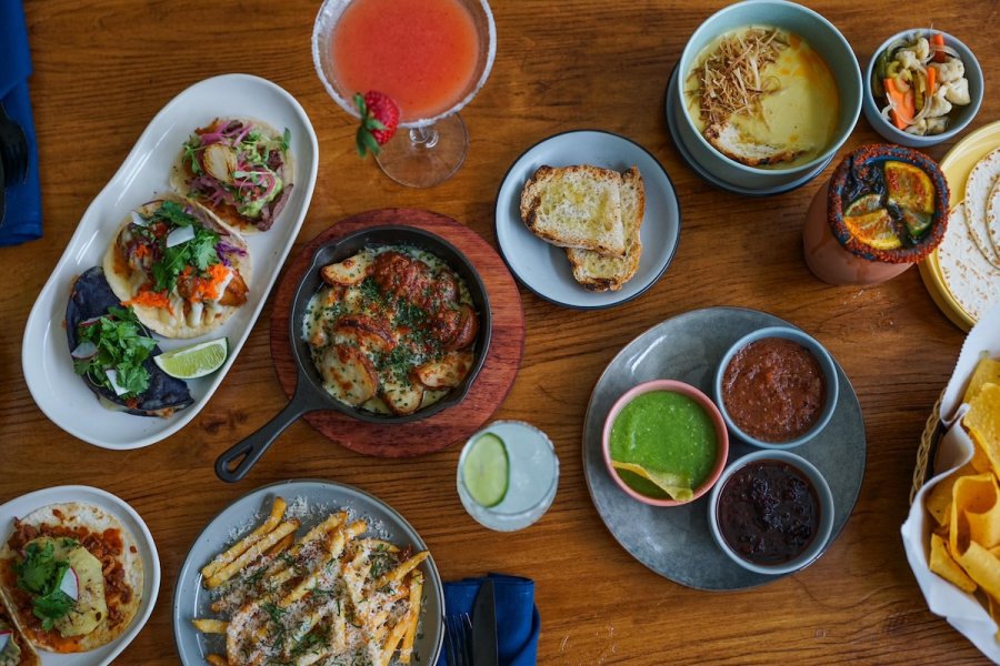 A wooden table set with various Mexican dishes, including tacos, cheese dip, sauces, fries, bread, cocktails, and chips with salsa—a vibrant scene often found in new restaurants Memphis food lovers adore.