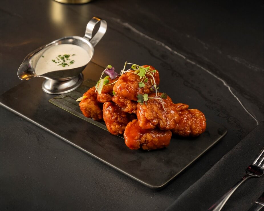 A serving of saucy boneless chicken wings garnished with microgreens on a black plate, accompanied by a small metal container of dipping sauce—a flavorful highlight among new restaurants in Memphis dining.