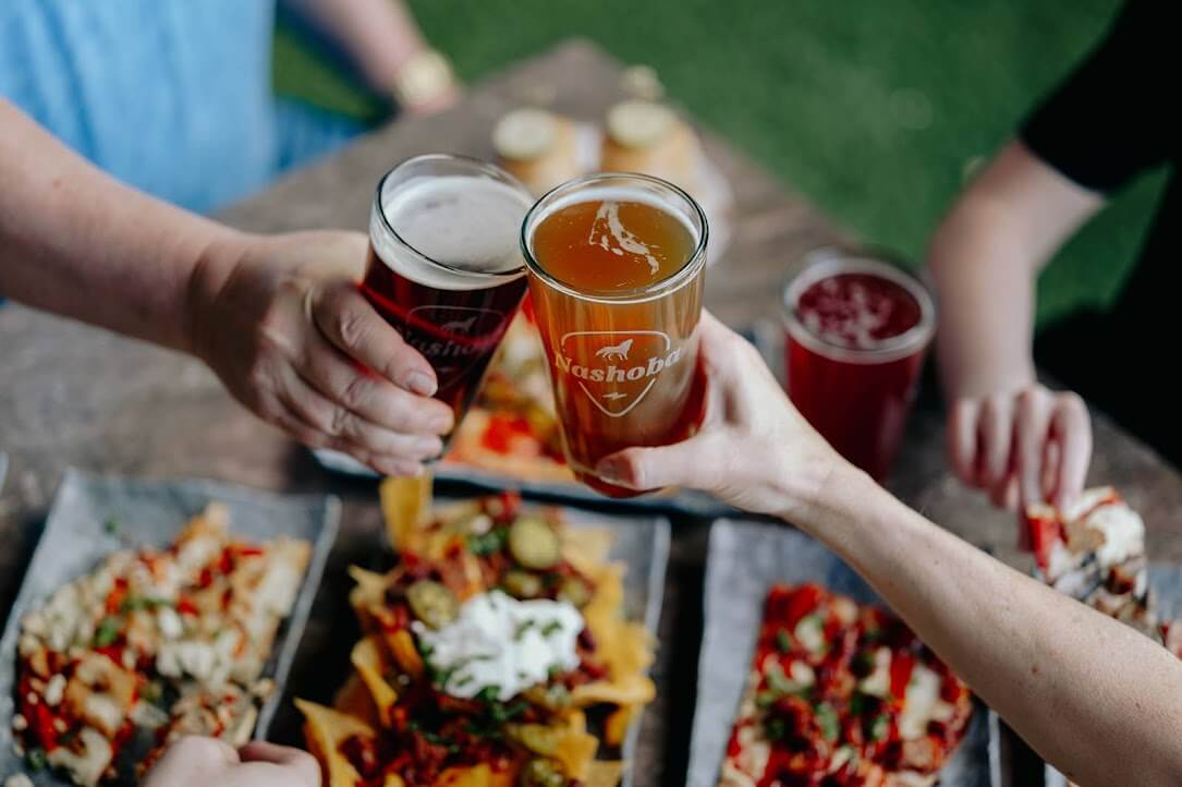 Two people clink glasses of beer over a table filled with loaded nachos and other food, enjoying the lively atmosphere of Memphis restaurants with friends seated around them.