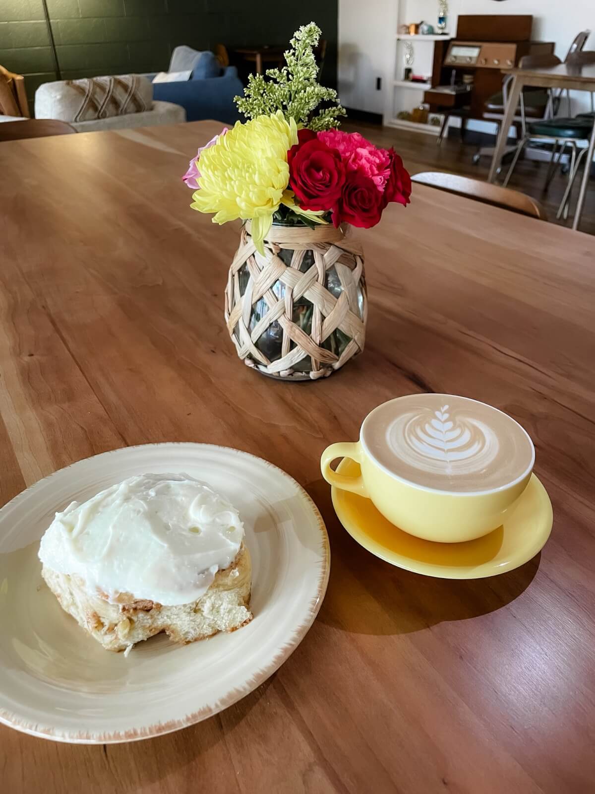 A cinnamon roll with icing on a plate and a cup of latte with latte art in a yellow cup sit on a wooden table next to a vase of colorful flowers, offering a cozy spot to enjoy Memphis dining.