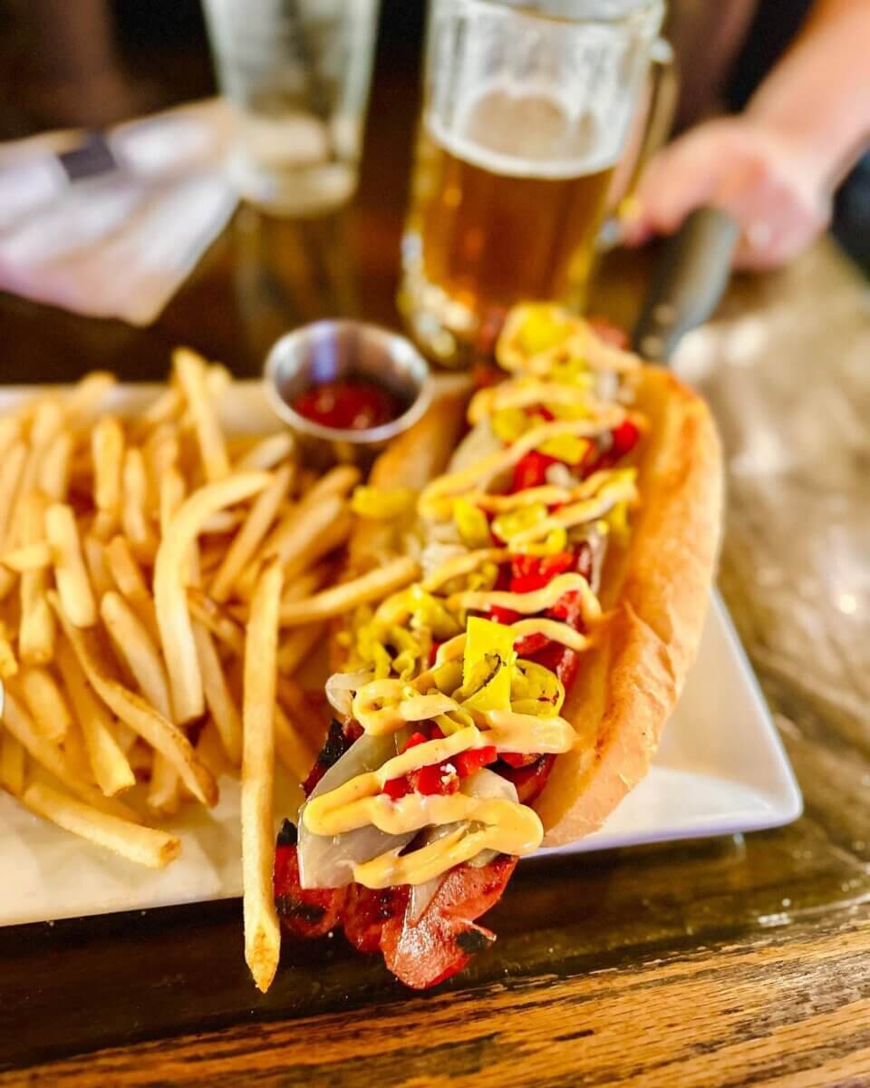 A grilled hot dog in a bun with mustard, relish, and onions, served with French fries, ketchup, and a glass of beer on a wooden table—perfect for enjoying at restaurants open on July 4th.