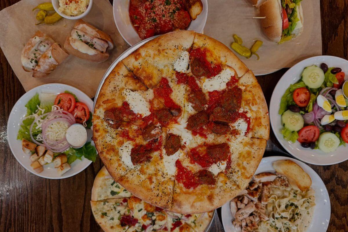 A table with a large pizza in the center, surrounded by salads, pasta, sandwiches, and various Italian dishes on white plates.