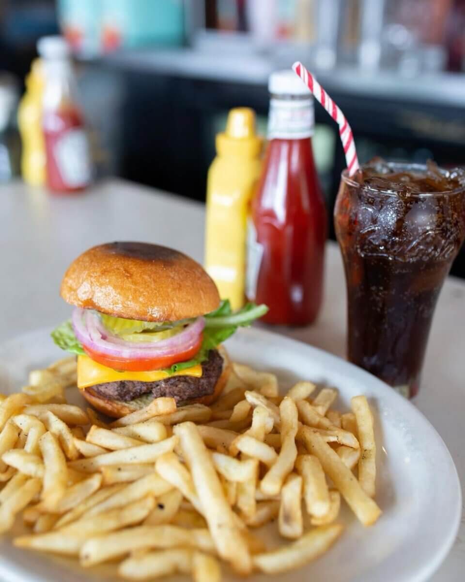 A plate with a cheeseburger topped with lettuce, tomato, and onion, served with French fries, sits next to a glass of soda—perfect for enjoying at restaurants open on July 4th, with ketchup and mustard bottles in the background.