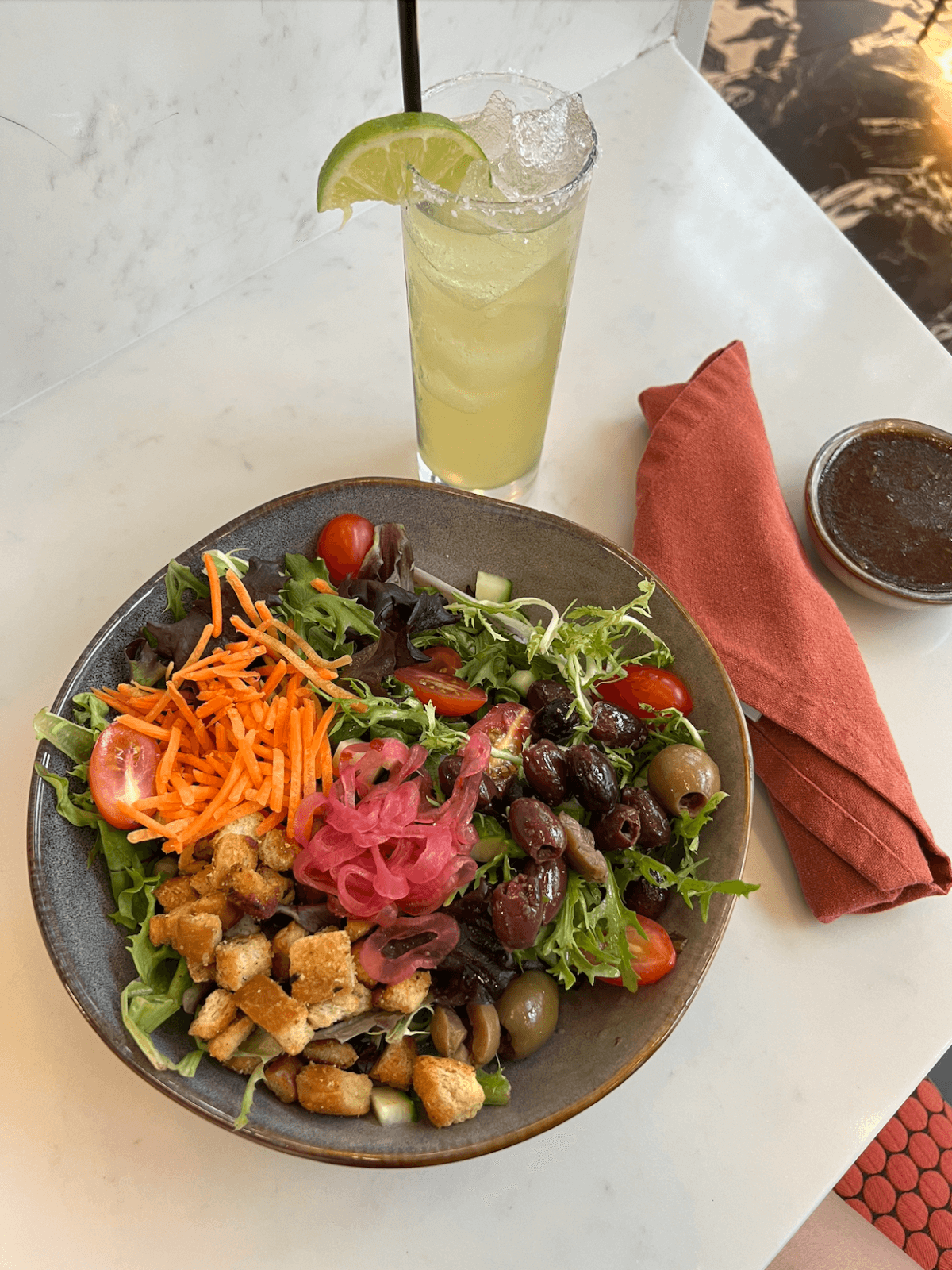 A bowl of mixed salad with croutons, olives, cherry tomatoes, shredded carrots, and pickled onions sits next to a drink with lime and an orange napkin on a white table—perfect for those seeking plant-based Memphis flavors.
