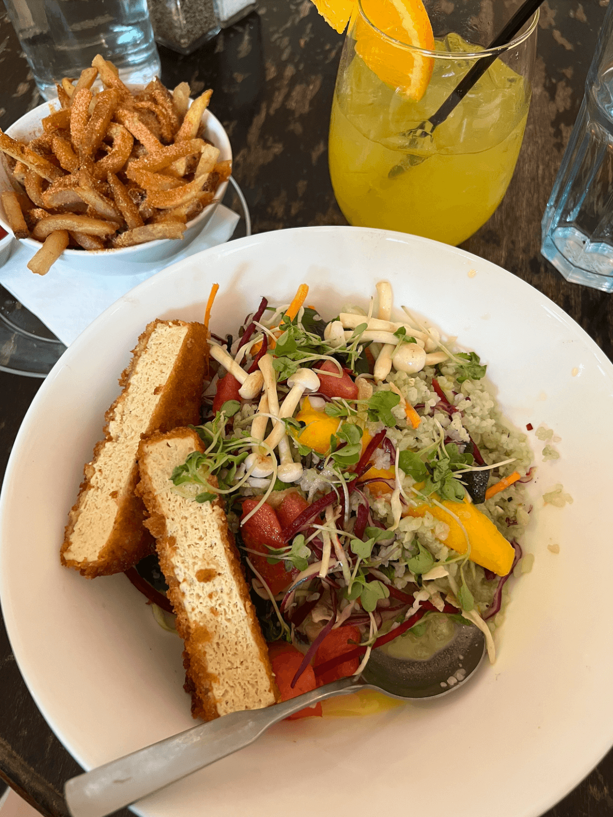 A Veg-Friendly bowl with sliced tofu, microgreens, vegetables, and grains, served alongside seasoned fries and a glass of orange drink garnished with an orange slice.