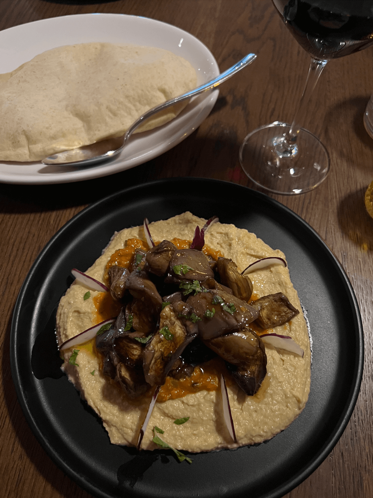 A plate of vegan hummus topped with roasted eggplant and herbs, served with flatbread, a spoon, and a glass of red wine on a wooden table—perfect for enjoying plant-based dining in Memphis.