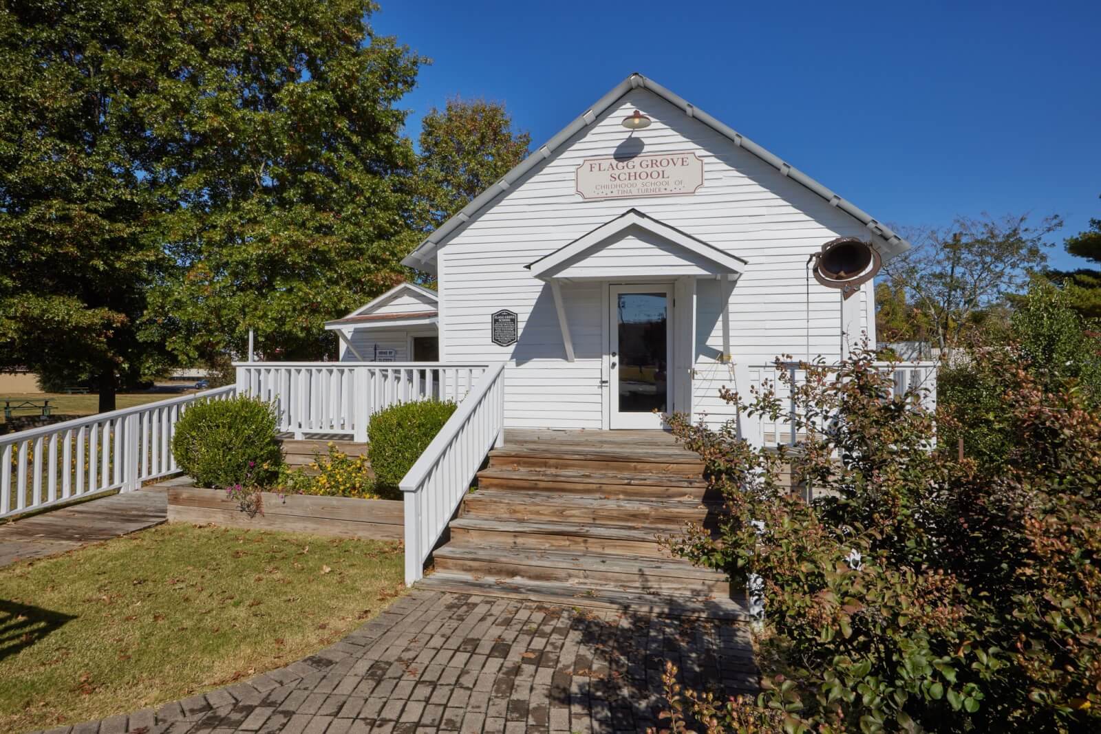 A small white wooden building with a front porch, labeled “Plum Grove School,” sits under a clear blue sky surrounded by greenery—an inviting spot for weekend getaways near Memphis.