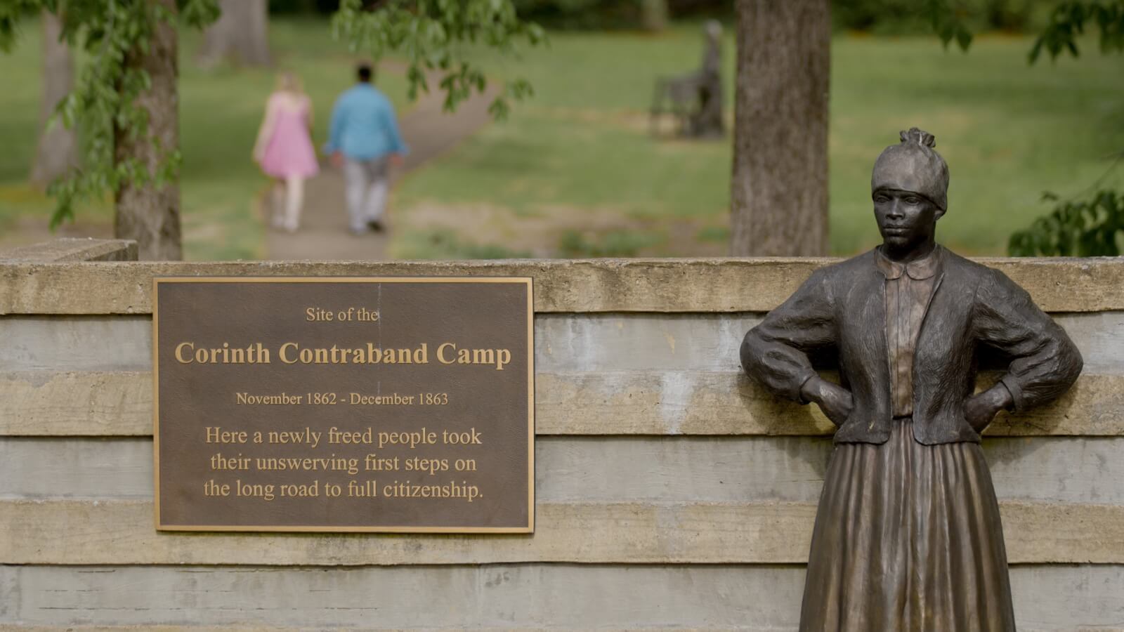 Bronze statue of a woman stands next to a plaque for the Corinth Contraband Camp; two people walk away on a path in the background—a meaningful stop on Weekend Getaways from Memphis.