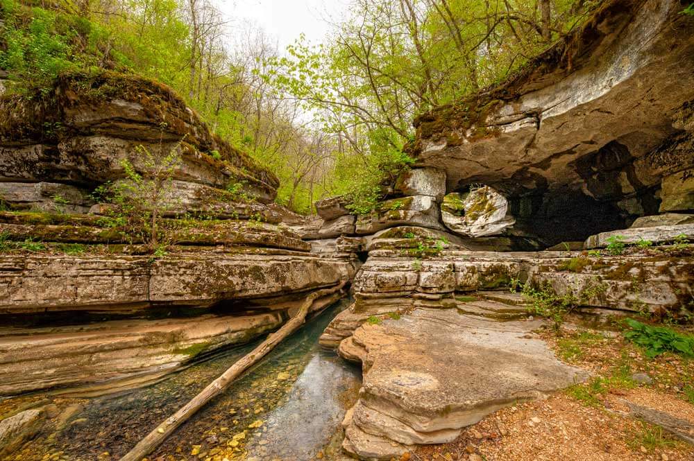 A rocky gorge with layered stone formations, a small creek, and green foliage surrounding the area under a cloudy sky—perfect for weekend getaways from Memphis.