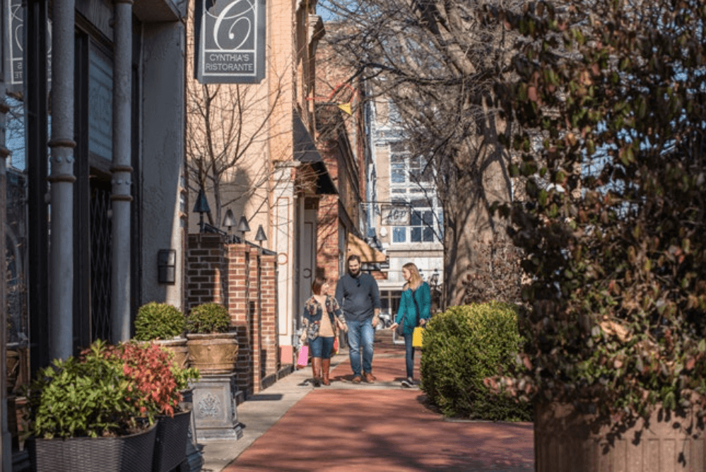 A family walks along a brick sidewalk past shops and plants on a sunny day in Memphis, with a sign for Cynthia’s Ristorante visible overhead—perfect scenery for spontaneous weekend getaways.