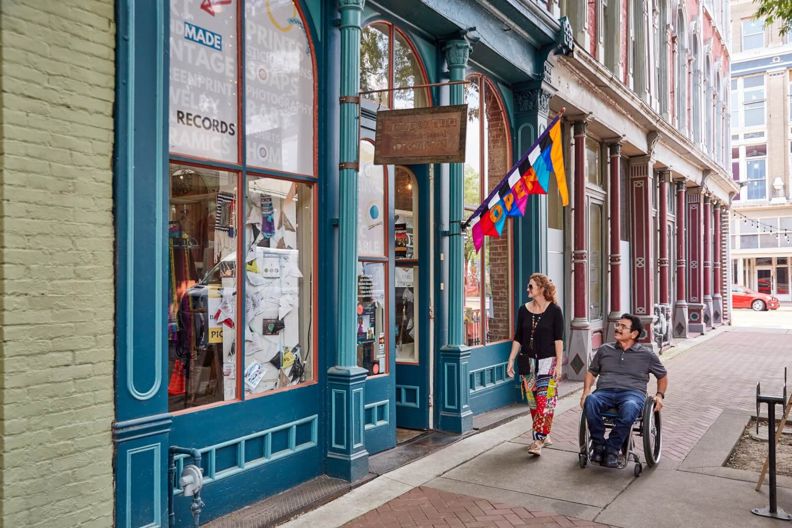Two people, one using a wheelchair, move along a Memphis sidewalk past shops with large windows and an "OPEN" flag—a welcoming scene for those seeking last minute getaways or weekend getaways in the city.