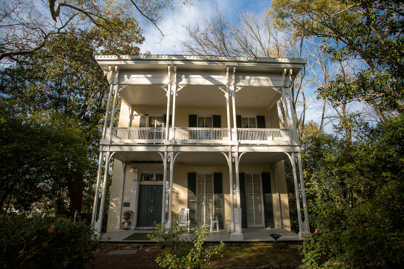 A two-story historic house with tall white columns, a double balcony, and many doors and windows, surrounded by trees and greenery—perfect for weekend getaways just outside Memphis.