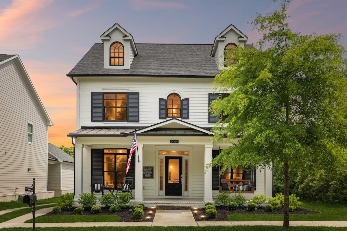 This storybook cottage is a stunning two-story white house with black shutters, front porch, and an American flag. Beautifully landscaped yard, captured perfectly at sunset.