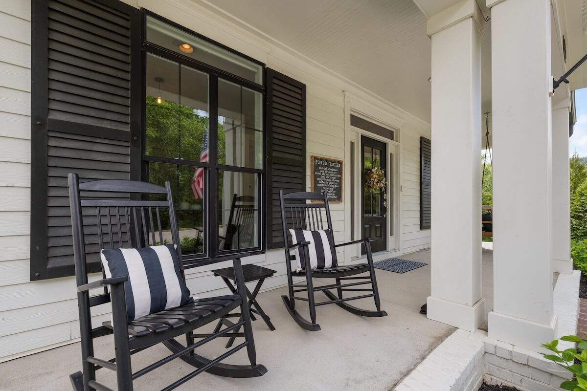 Two black rocking chairs with striped pillows sit on the covered porch of this storybook home, featuring white siding, black shutters, and a black front door. An American flag is visible through the window of this charming Franklin Cottage.
