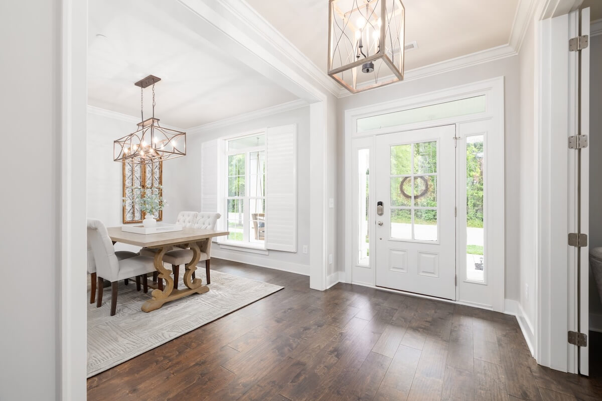 This storybook cottage features a bright entryway with wood floors leading to a dining room with a wooden table, upholstered chairs, large windows, and modern light fixtures.