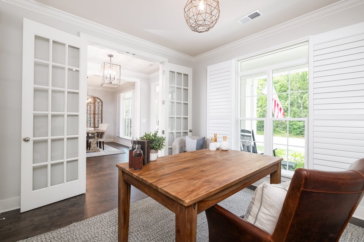 A bright room in the Franklin Cottage with a wooden table, leather chair, and decorative items features large windows and French doors opening to another room. An American flag is visible outside. Just Listed!.