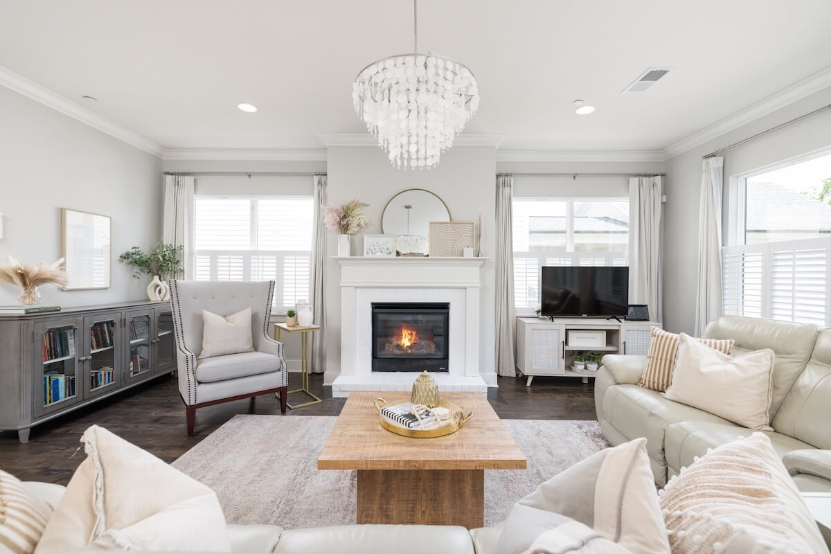 Bright, modern living room in the Just Listed Franklin Cottage features a fireplace, neutral sofas, armchair, wooden coffee table, bookshelves, and TV. A chandelier graces the ceiling as light streams through large windows.