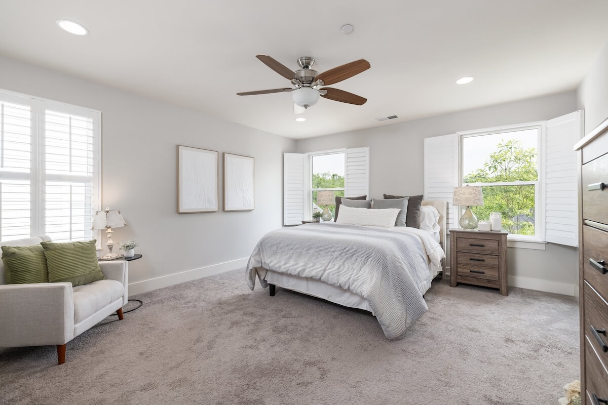 Bright, neutral-toned bedroom at Franklin Cottage with double bed, armchair, wooden nightstands, ceiling fan, and open shuttered windows letting in natural light. Just listed and ready for your storybook cottage dreams!.