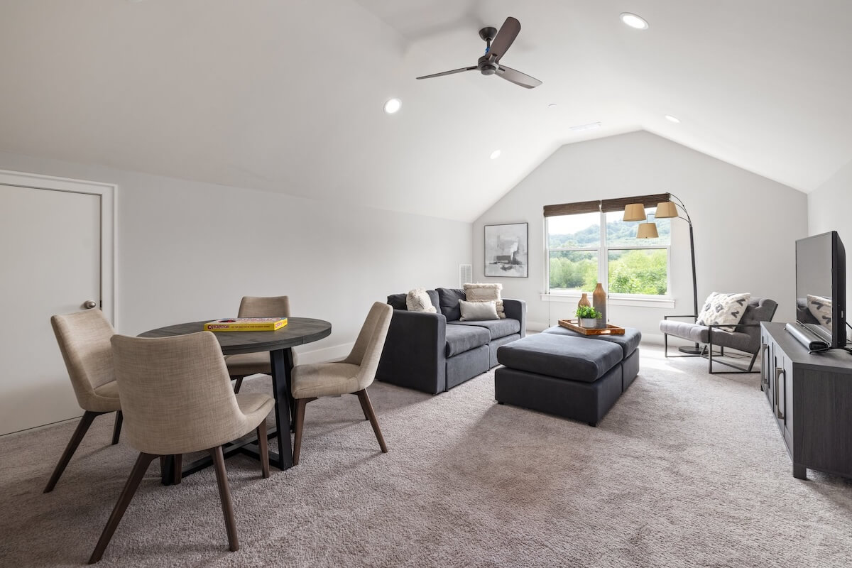 A modern living room in Franklin Cottage features a round dining table, four chairs, two gray sofas, an ottoman, a TV on a stand, and a window with views of greenery—perfect for those seeking storybook cottage homes for sale.