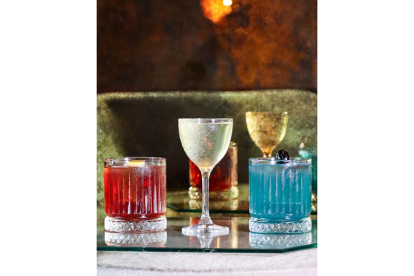 Three cocktails in ornate glasses—one red, one clear, and one blue—are artfully arranged on a reflective surface at the Four Walls Bandista Pop-up, set against a blurred, dimly lit background.