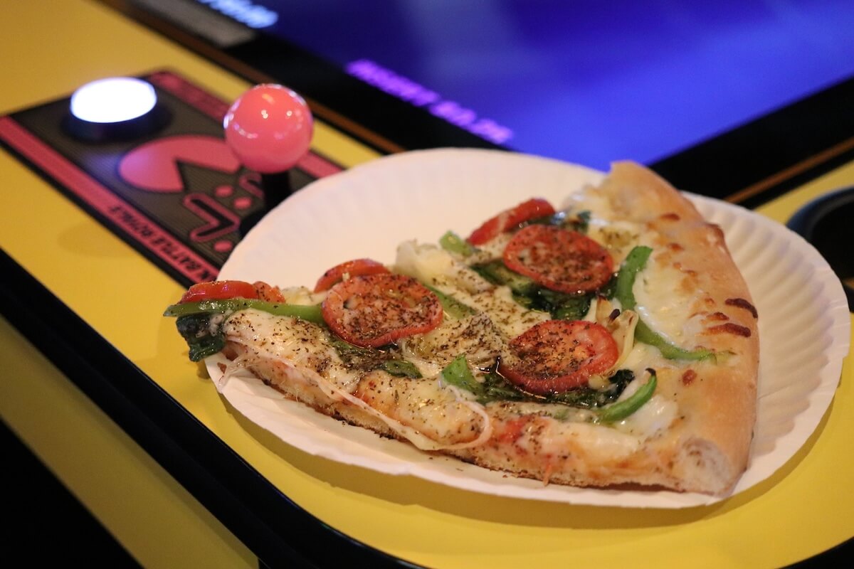 A slice of vegetable pizza on a paper plate rests on a yellow arcade game console with a pink joystick and a white button.