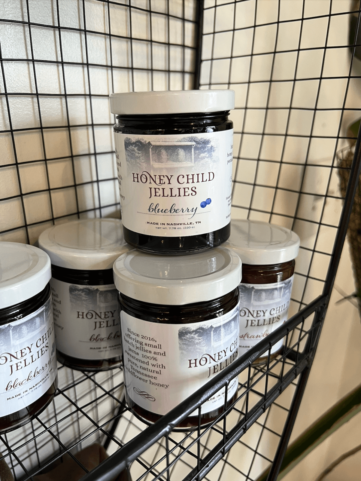 June FINDS: Jars of Honey Child Jellies blueberry jam with white lids are stacked on black wire shelves against a white wall in Nashville.