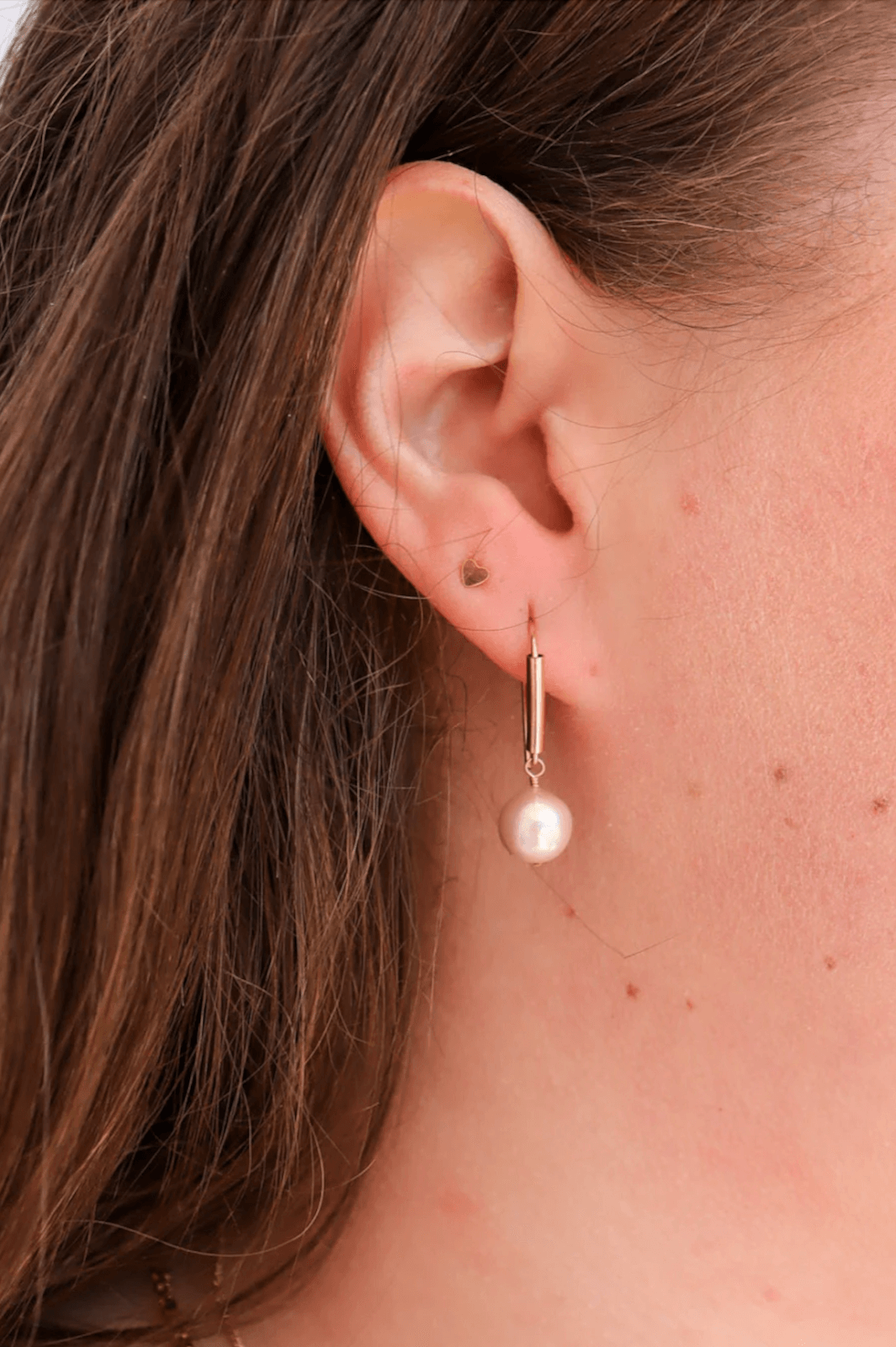 Close-up of a woman's ear with a small gold stud earring in the upper lobe and a gold drop earring with a pearl in the lower lobe. Brown hair partially visible—a stylish June FINDS look inspired by Nashville.