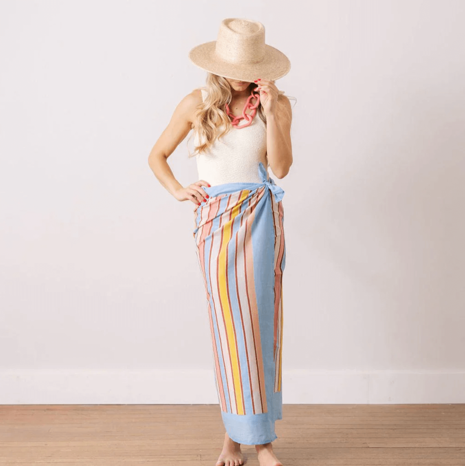 A person wearing a wide-brimmed hat, white sleeveless top, red necklace, and a long striped wrap skirt stands against a plain white wall and wooden floor—an effortless June look inspired by Nashville FINDS.